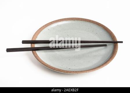 A pair of oriental black wooden chopsticks rests on a textured stoneware plate and set on plain white background. Stock Photo