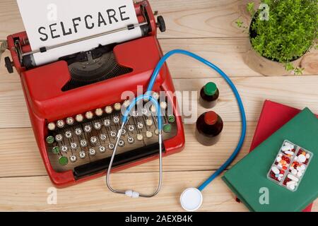Prescription medicine or medical diagnosis - doctor workplace with blue stethoscope, pills, red typewriter with text Selfcare on wooden table Stock Photo