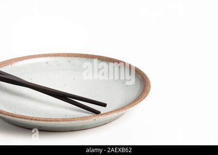 A pair of oriental black wooden chopsticks rests on a textured stoneware plate and set on plain white background. Stock Photo