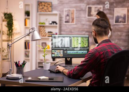 Back view of creative filmmaker working on a movie on laptop. Girlfriend in the background. Stock Photo