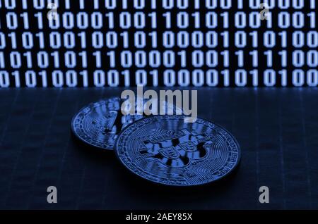 A pair of bitcoins lies on a cardboard surface on the background of a monitor depicting a binary code of bright zeros and one units on a black backgro Stock Photo