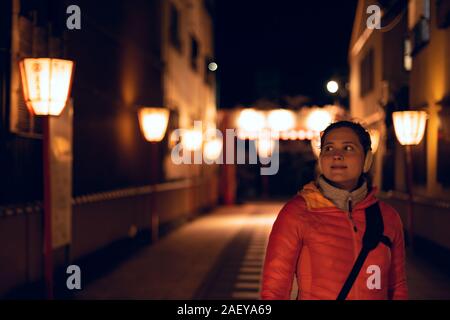 Kyoto, Japan narrow alley dark street in Gion district at night with young happy woman standing looking at illuminated row of red lanterns Stock Photo