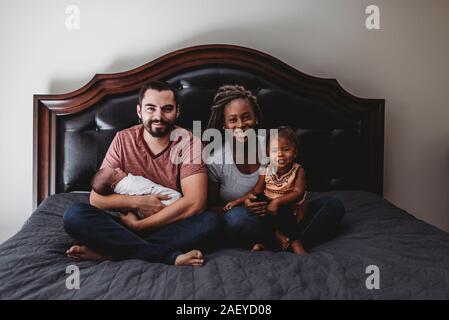 Happy multiracial family on bed with dad & infant and mom & 2yr old Stock Photo