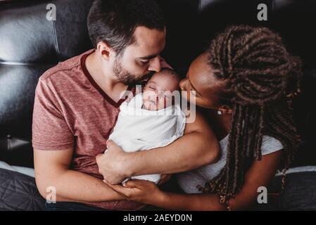 Multiracial parents kissing newborn baby wrapped in white blanket