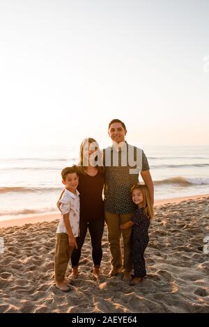 Family of Four Smiling at Camera, Embracing on Beach at Sunset Stock Photo