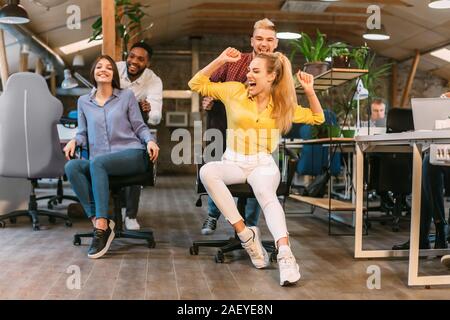 Positive colleagues having fun in office, enjoying office chair race and competing Stock Photo