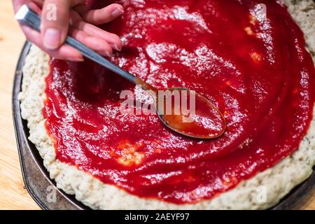 Baking pan with uncooked pizza dough crust before baking made with gluten free flour and woman hands adding tomato sauce with spoon flat top view Stock Photo