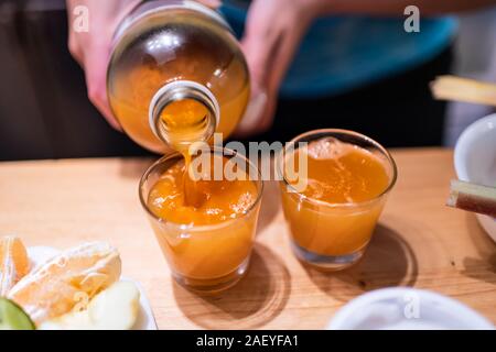 Woman person serving pouring kombucha fermented tea into two juice shot glasses on wooden table from bottle Stock Photo