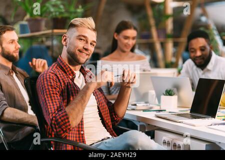 IT development. Positive man smiling to camera at workplace, sitting in coworking office with colleagues Stock Photo