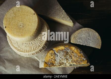 Different homemade cheeses on a dark wooden background. Pieces and cheese heads of Gorgonzola, Asiago, Maasdam, Gauda, Paramezan and Edam. Goat and cow cheese on baking paper. Handwork, Flat lay. Stock Photo