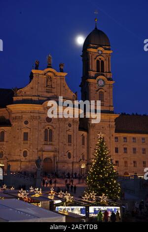 Einsiedler Weihnachtsmarkt (Christmas market) in front of the monastery Einsiedeln at dawn with a christmas tree, booths and the moon shining above. Stock Photo