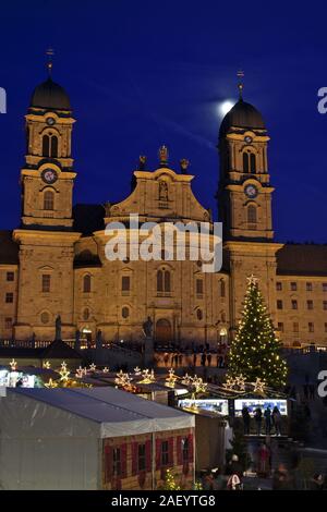 Einsiedler Weihnachtsmarkt (Christmas market) in front of the monastery Einsiedeln at dawn with a christmas tree, booths and the moon shining above. Stock Photo