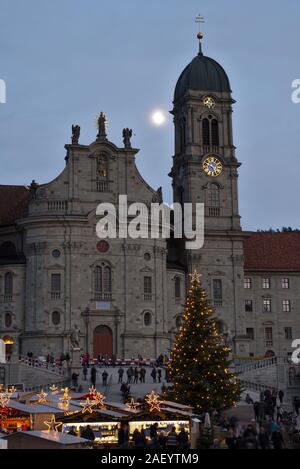 Einsiedler Weihnachtsmarkt (Christmas market) in front of the monastery Einsiedeln at dawn with a christmas tree, booths and the moon shining above. Stock Photo