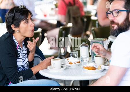 New Orleans, USA - April 23, 2018: Couple man and woman sitting at table at famous Cafe Du Monde coffee shop restaurant drinking chicory eating beigne