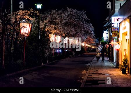 Kyoto, Japan - April 9, 2019: Red paper lanterns lamps glowing with cherry blossom flower trees along Kamo river during Hanami festival and couple wal Stock Photo