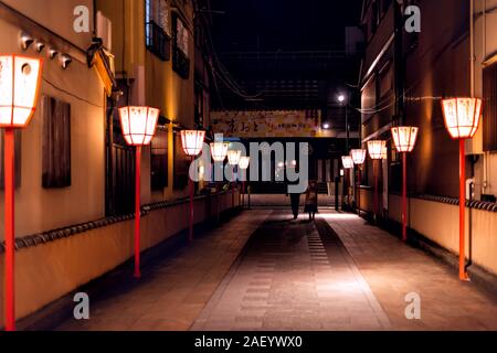 Kyoto, Japan - April 9, 2019: Narrow alley dark street in Gion district at night with couple people walking and illuminated row of red lanterns Stock Photo