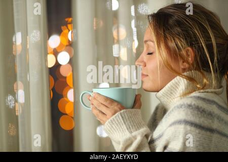 Side view portrait of a woman smelling coffee beside a window standing in the night at home Stock Photo