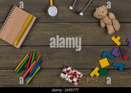Children's doctor or pediatrician concept - blank notepad, pills, yellow stethoscope, colorful wooden jigsaw puzzles, Teddy bear toy, crayons on woode Stock Photo
