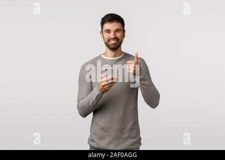 Nice, thanks for recommendation. Cheerful handsome bearded man in grey sweater holding smartphone, make thumb-up gesture, like app, advertise Stock Photo