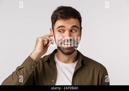 Hello, I am listening. Cheerful, friendly-looking handsome bearded caucasian man talking on phone, answer with tapping wireless earphone, wearing Stock Photo