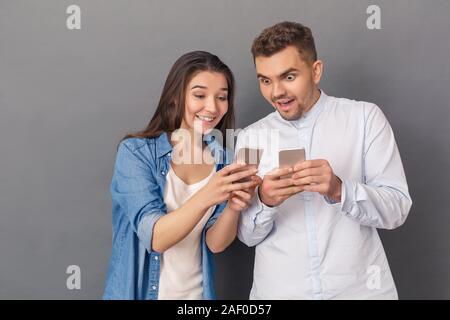 Using Devices. Young couple studio standing isolated on grey showing each other media on smartphone smiling excited Stock Photo