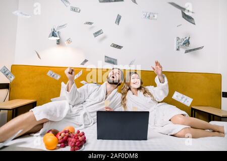 Happy young couple in white bathrobes throwing currency notes in air, while lying on the bed in modern bedroom or hotel. Laptop, fruits on the bed. Stock Photo