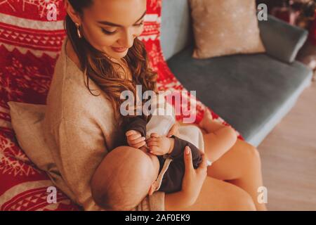 Mother feeds her baby with a bottle, artificial feeding, New Year's interior, mother sits on a sofa Stock Photo
