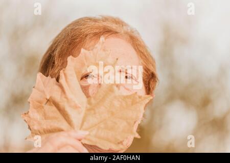 The senior woman looking through the heart-shaped hole in the maple leaf. Autumn concept. Selective focus Stock Photo
