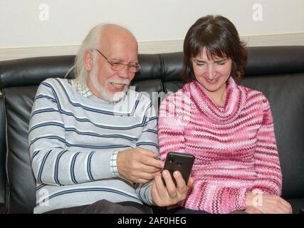 Laughing senior man is demonstrating something funny in his smartphone to mature dark-haired woman that sitting beside him on black leather sofa. Stock Photo