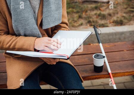 Cropped view of blind man reading book with braille font beside walking stick and thermo mug on bench Stock Photo