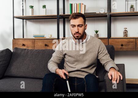 Visually impaired man holding walking stick while sitting on sofa in living room Stock Photo