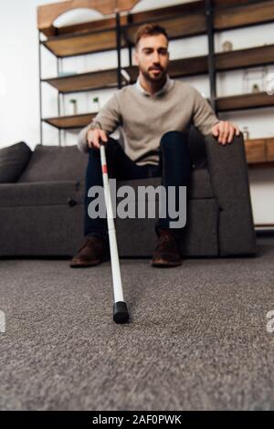 Selective focus of visually impaired man holding walking stick while sitting on sofa in living room Stock Photo