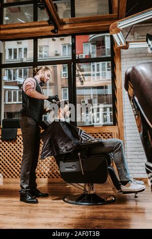 side view of handsome barber styling hair of man in modern barbershop Stock Photo