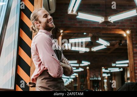 happy barber in apron standing with crossed arms in barbershop Stock Photo