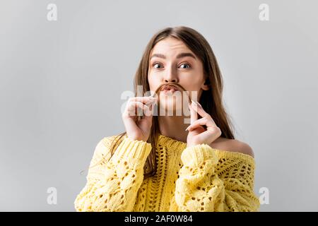 tricky girl in yellow sweater holding hair above lip like mustache isolated on grey Stock Photo