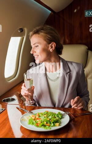 happy businesswoman with champagne glass near tasty salad in private jet Stock Photo