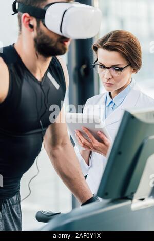 sportsman running on treadmill near attractive doctor during endurance test in gym Stock Photo