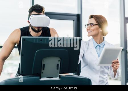 sportsman in virtual reality headset running on treadmill near smiling doctor during endurance test Stock Photo