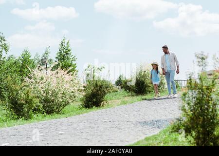 handsome father and daughter walking near green plants and holding hands Stock Photo
