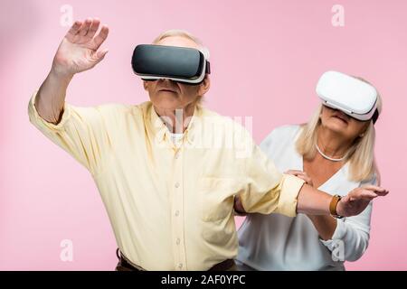 retired man gesturing while wearing virtual reality headset near wife on pink Stock Photo