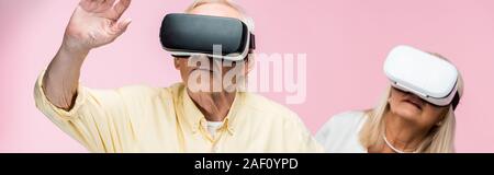 panoramic shot of retired man gesturing while wearing virtual reality headset near wife on pink Stock Photo