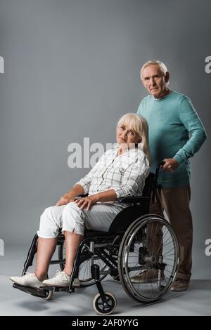 upset man standing near disabled retired wife sitting in wheelchair on grey Stock Photo