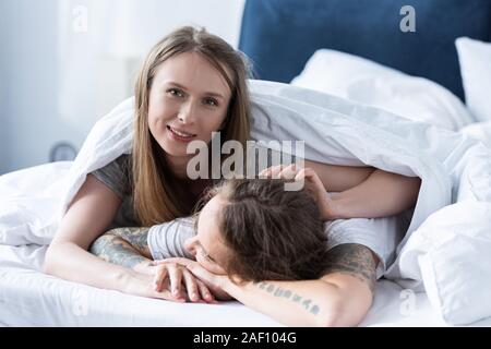two smiling lesbians embracing while lying under blanket in bed Stock Photo