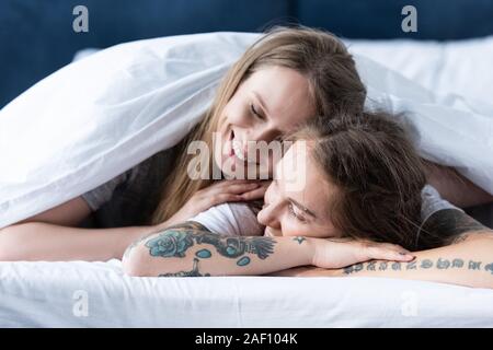 two smiling lesbians embracing while lying under blanket in bed Stock Photo
