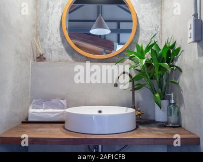 Bangkok, Thailand - December 1, 2019: Loft style bathroom interior. Round mirror on concrete wall with brass faucet, modern white sink basin, green le Stock Photo