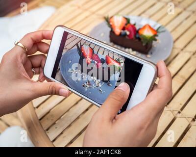 Woman's hand taking a photo of delicious chocolate tart on ceramic dish on bamboo table background by smartphone in cafe. Smartphone food photography Stock Photo