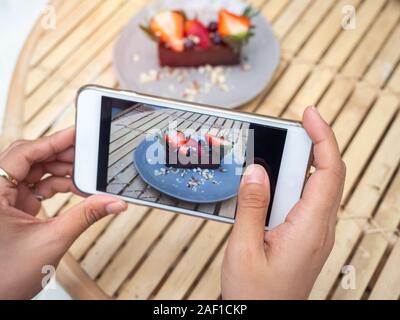 Woman's hand taking a photo of delicious chocolate tart on ceramic dish on bamboo table background by smartphone in cafe. Smartphone food photography Stock Photo