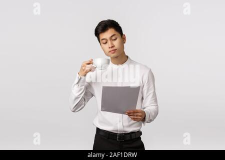 Business, corporate concept. Handsome asian businessman drinking coffee from cup, look at documents, reading report, studying charts as working in Stock Photo