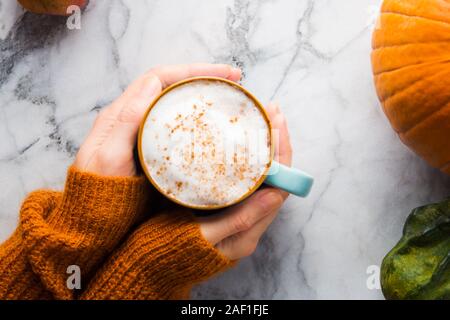 Autumn moody background with mug of latte coffee and pumpkins on marble table. Flat lay in fall colors. Female hands in cozy sweater Stock Photo