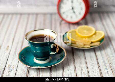 Green cup of hot tea, saucer with lemons and candied ginger on a wooden table with a red alarm clock in the background Stock Photo